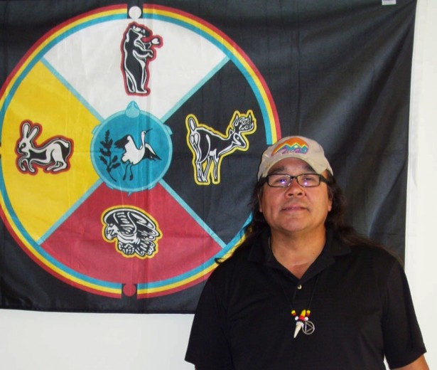 Manidoogekek, standing in front of the Ojibwe tribal flag, which hangs on the wall of his office at Native American Lifelines in West Roxbury, Massachusetts. Some of the programs offered there include smoking cessation, one-on-one counseling, group counseling, HIV prevention, behavioral health and treatments for substance abuse. Manidoogekek proudly reports that “plans for Red Road Recovery meetings and ceremony are in the works.” (By Donna Laurent Caruso)